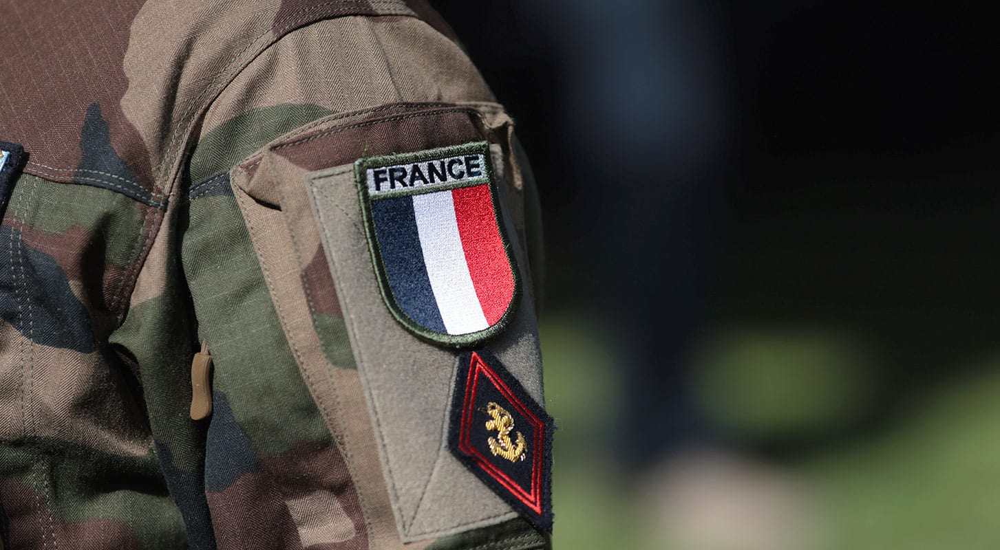 A soldier in uniform with a French flag patch on his or her jacket stands at attention.